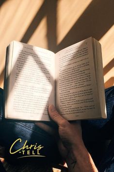 a person is holding an open book in their hand while sitting on the floor with sunlight streaming through them