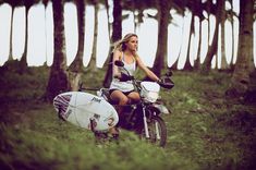 a woman riding on the back of a motorcycle next to a white surfboard with trees in the background