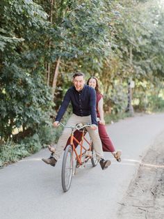 a man and woman riding on the back of an orange bicycle down a road with trees in the background