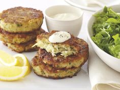 two crab cakes are next to a bowl of salad and a lemon wedge on the side