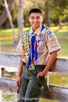 a boy in scout uniform leaning on a fence with his hand on his hip and smiling at the camera