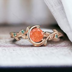 a gold ring with an orange stone surrounded by green stones and leaves on the side