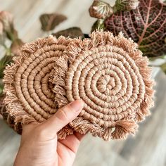 a hand holding up a woven piece of art with leaves in the background and a person's hand reaching for it