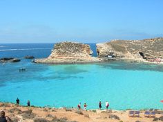 people are standing on the beach near some rocks and clear blue water, while others swim in the ocean