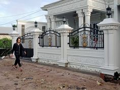 a woman walking down a dirt road past a white building with black iron gates on it