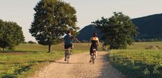 two people riding bikes down a dirt road in the country side with trees and hills behind them