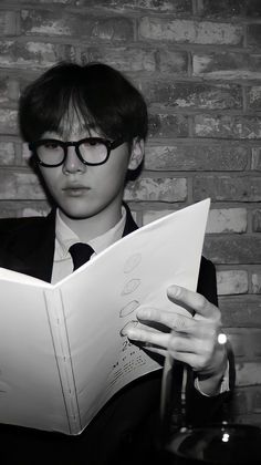 a man wearing glasses reading a book in front of a brick wall