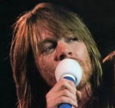 a man with long hair holding a white and blue object in his mouth while singing into a microphone