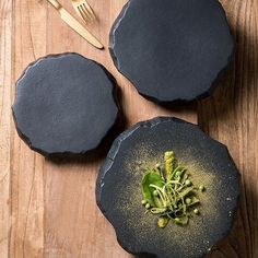 three black plates with food in them on top of a wooden table next to utensils