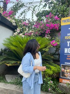 a woman standing in front of a blue sign next to purple and white flowered bushes