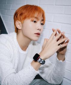 a young man with red hair wearing a white sweater and black wrist watch sitting in front of a brick wall