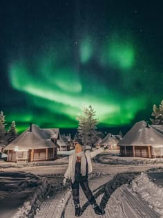 a woman is standing in the snow with an aurora bore above her and houses behind her