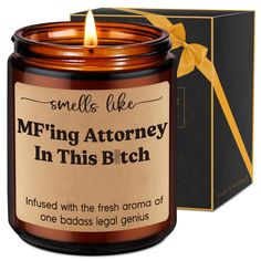 a candle that is in front of a box with a ribbon on it and the label reads, smells like m f ing atteney in this b t