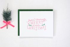 a christmas card next to a pine tree branch with pink ribbon and merry message on it