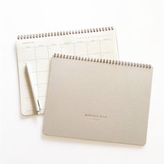 two spiral bound notebooks sitting next to each other on a white surface with a pen