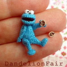 a small blue knitted cookie monster brooch sits on someone's finger and looks like he is holding something in his hand