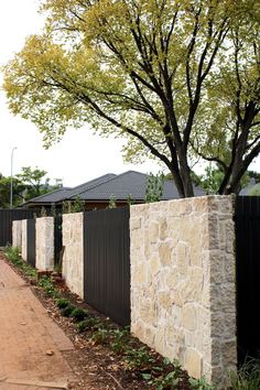 a stone wall next to a tree in front of a black fence and trees with green leaves