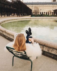 a woman is sitting on a bench in front of a fountain and looking at the water