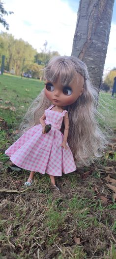 a doll is sitting in the grass next to a tree and wearing a pink dress