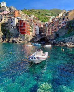 boats floating on the water in front of colorful buildings and cliffs, with people swimming nearby