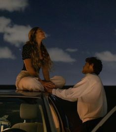 a woman sitting on top of a car next to a man who is holding his hand out