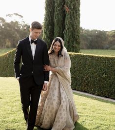 a man and woman dressed in formal wear walking through the grass with bushes behind them