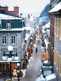 a city street is covered in snow and decorated with christmas lights on the buildings, along with people walking down it