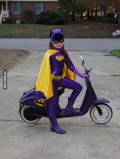 a woman dressed as batgirl sitting on a scooter in the middle of a driveway