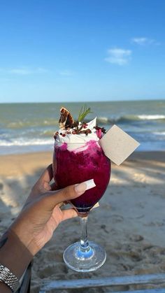 a person holding up a wine glass with food in it on the beach next to the ocean