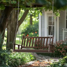 a wooden swing hanging from a tree in front of a house with flowers and trees around it