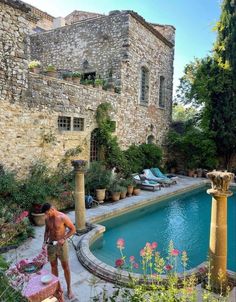 a man standing in front of a pool surrounded by plants and flowers, next to a stone building