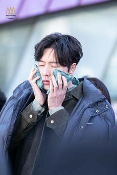 a man talking on a cell phone while wearing a blue jacket and black coat with his hands to his face