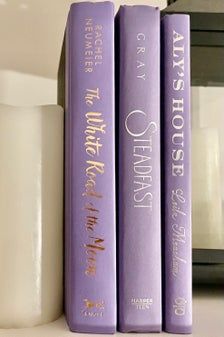 three purple books sitting next to each other on a shelf near a white cup and candle