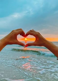 two hands making a heart shape with the sun setting in the distance over the ocean