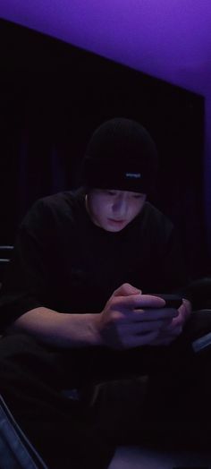 a young man sitting in the dark looking at his cell phone while wearing a beanie