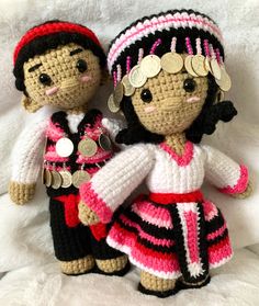 two crocheted dolls are posed on a white blanket, one is wearing a pink and black dress