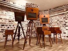 four wooden chairs and two televisions on stands in front of a brick wall with wood flooring