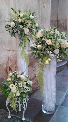 two tall vases with flowers and greenery in them on a stone floor next to a brick wall