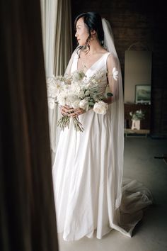 a woman standing in front of a window holding a bouquet of flowers and wearing a wedding dress