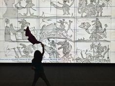 a woman walking past a large screen with drawings on it's walls and people in the background