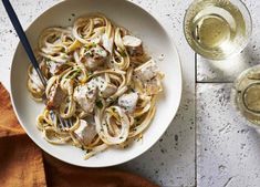 a white bowl filled with pasta and meat on top of a table next to two glasses of wine