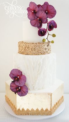 a three tiered white cake with purple flowers on top and gold trimmings