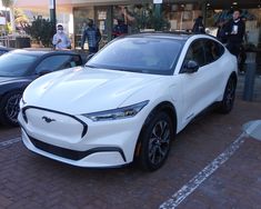 a white electric car parked in front of a building with people standing around and looking at it