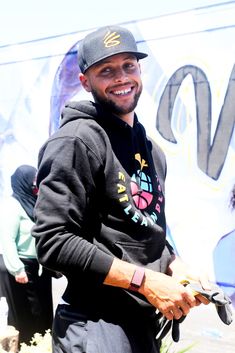 a man in a black hoodie and cap smiles at the camera while holding a skateboard