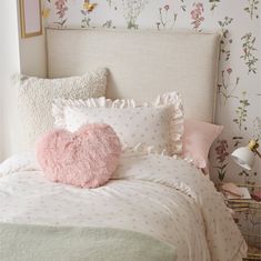 a white bed with pink pillows and a heart shaped pillow on top of it in front of a floral wallpaper