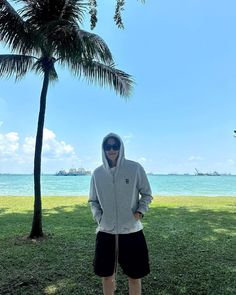 a man standing in front of a palm tree on the grass near the ocean with his hands in his pockets