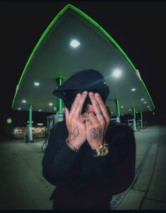 a man in a black suit and hat covering his face with both hands while standing next to a gas station