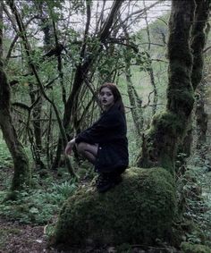 a woman sitting on top of a moss covered rock in the woods with trees behind her