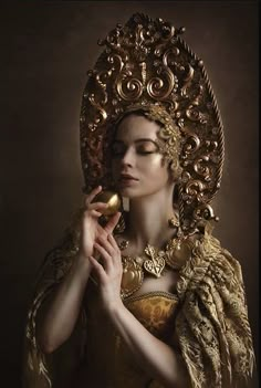 a woman in an ornate gold dress holding a golden object up to her face with both hands