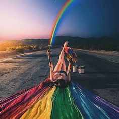 a woman laying on the ground with a rainbow kite in front of her and a rainbow painted sky behind her